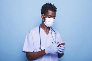 African american medical assistant in blue scrubs grasping smartphone in hand. Male healthcare specialist wearing face mask and gloves for covid 19 protection while having a mobile device. photo