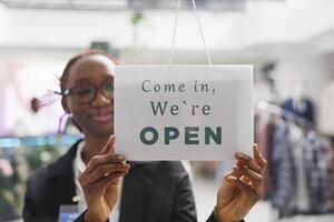 Fashion boutique shop smiling employee hanging we are open sign on store front door. Young african american clothing mall department manager holding signboard with come in message photo