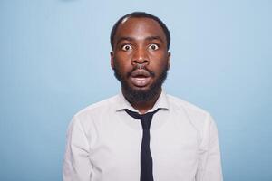 Stunned businessman stands in front of an unbelievable situation on a blue background, his mouth hanging open in shock. Surprised black individual with wide eyes and a jaw drop. photo