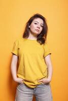 Confident woman with hands in pocket, brown hair, and casual clothing stands against an isolated orange background. Her expressive eyes and friendly demeanor are captivating. photo