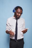 joven negro hombre con inalámbrico auriculares utilizando teléfono inteligente a vistazo social medios de comunicación mientras participación un calentar beber. en su romper, oficina trabajador escucha a en línea música mientras Bebiendo un taza de café. foto