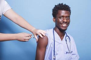 masculino médico enfermero vistiendo azul matorrales consiguiendo vacuna inyección con jeringuilla en estudio. africano americano médico con estetoscopio, consiguiendo vacunado para coronavirus inmunidad. foto