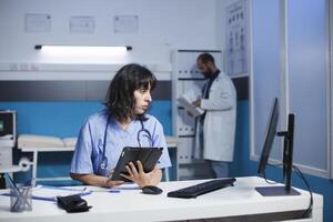 hembra enfermero usos un inteligente tableta y computadora para médico información y chequeos. mientras examinando paciente datos en escritorio ordenador personal, cuidado de la salud profesional sostiene un artilugio con un toque pantalla. foto