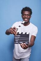 African american filmmaker holding open clapper in hands looking confident with a smile on his face. Black movie producer with clapperboard standing proud in front of isolated blue background. photo