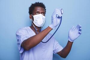 Male healthcare nurse is holding stethoscope to examine cardiology patient heartbeat. African American man wearing gloves and face mask as cardiologist fights coronavirus epidemic. photo