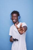 African American man wearing headphones and carrying controller gesture peace sign towards camera. Black person standing in front of isolated backdrop makes happy gesture with their hand. photo