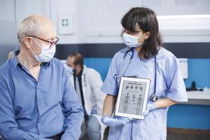 Healthcare specialist describes human skeleton image on tablet to senior patient with face mask at medical exam. Female physician grasps device with osteopathy diagnosis, showing bones examination. photo