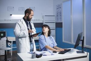 Caucasian doctor and female healthcare worker collaborate in a clinic office. They discuss digital medical images on a tablet and desktop computer. Medical equipment is visible. photo