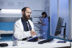 imagen vitrinas un dedicado caucásico médico comprobación y analizando su notas a el clínica oficina escritorio. joven hombre vistiendo un blanco laboratorio Saco preparando para médico consultas con pacientes foto