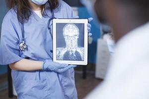 en médico oficina, médico usos tableta a analizar un escanear de un del paciente cráneo. selectivo atención de enfermero en azul matorrales participación un dispositivo con un radiografía imagen. avanzado tecnología y experto cuidado. foto