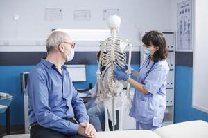 Nurse wearing face mask is describing bones in human skeleton to elderly guy in cabinet. They are also discussing osteopathy anatomy and spinal cord discomfort. Doctor doing a physical assessment. photo