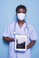 Male nurse looking at camera while holding a tablet with blank screen, against blue background. Demonstrating healthcare expertise, black man wears scrubs, face mask, gloves, and stethoscope. photo