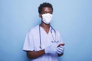 Close-up of nurse wearing face mask, holding smartphone for healthcare information during pandemic. African American medical professional stands confidently against blue background with mobile device. photo