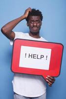 Bewildered african american individual holding voice bubble with text message asking for help. Portrait of black guy with hand on his head is grasping a red cardboard sign. photo