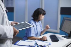 Close-up shot of medical professionals utilizing modern technology for discussing healthcare in a clinic. Selective focus shows a person holding a tablet and a female nurse using a computer. photo