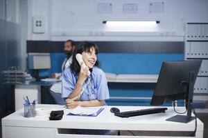 sonriente hembra facultativo sentado a el clínica oficina escritorio es hablando en el teléfono fijo caucásico enfermero en teléfono llamada mientras utilizando un escritorio computadora en un hospital habitación. foto