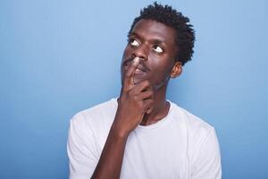 retrato de negro hombre lluvia de ideas y reflexionando mientras avaro su mentón. en estudio, enfocado africano americano chico con mano en rostro, pensando y preguntándose en frente de azul antecedentes. foto