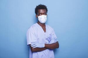 en estudio, enfermero con cruzado brazos, vistiendo cara máscara y guantes, mira a cámara. negro hombre trabajando como médico asistente soportes terminado aislado fondo, protegido en contra codicioso 19 pandemia. foto