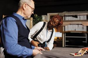 Helpful old master suitmaker and happy tailor apprentice cutting fabric sheet for upcoming stylish sartorial collection. Couturiers in atelier studio manufacturing refined garments photo