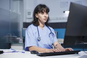 hembra facultativo en un moderno clínica oficina utilizando un teclado a tipo médico notas en un computadora. caucásico enfermero con un estetoscopio actualización paciente información en escritorio ordenador personal. foto