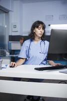 In clinic office, committed doctor using computer to analyze patient records. Female nurse checking patient information on a desktop computer in the hospital with focused attention. photo