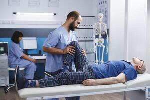 Physiotherapist assisting senior man with leg injury to recover at kinesiotherapy clinic. Nurse practitioner giving support to retired patient with knee pain and muscle pressure for recovery. photo