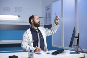 joven médico examinando un Connecticut escanear de paciente y tomando notas en su bloc. caucásico masculino cuidado de la salud profesional inspeccionando un cofre radiografía imagen de un individual. foto