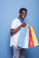 African American guy wearing sunglasses, holding shopping bags, and standing in front of an isolated blue background. Black man with wireless headset on his neck, carrying colorful parcels. photo