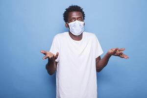 Clueless african american man wearing a face mask raises his shoulders in confusion. Standing against isolated backdrop is black guy staring at camera with an expression of uncertainty. photo