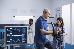 Nurse practitioner giving assistance to old man using stationary bike for physical activity and recovery. Medical assistant helping male patient with exercise bicycle for physiotherapy. photo