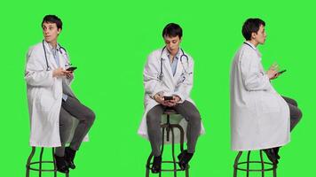 Woman specialist using smartphone social media apps in studio, sitting on a chair against greenscreen backdrop. Physician waiting for someone and browses online webpages, texting. Camera A. photo