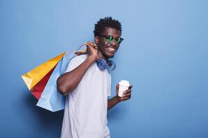alegre africano americano hombre participación un taza en felicidad después un compras juerga. un optimista masculino individual vistiendo Gafas de sol y auriculares, agarrando pantalones con productos desde negro viernes. foto
