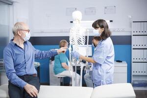 Senior man discussing with nurse about human skeleton for chiropractic care. Female physician points at spinal cord to explain diagnosis for osteopathy and recovery. Nurse doing bones presentation. photo