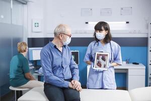 Senior man looking at heart illustration on tablet held by medical assistant. Female practitioner describing cardiovascular system and treatments to male elderly patient while wearing face masks. photo