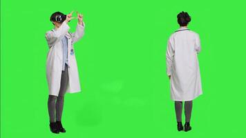 Woman medic works with 3d virtual reality headset in studio, standing against greenscreen backdrop. General practitioner uses vr glasses with interactive vision tool, futuristic exam. Camera A. photo