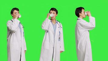 Woman physician drinking a cup of coffee against greenscreen backdrop, taking a break from medical work. Specialist serving caffeine refreshment in studio, wearing a white coat. Camera B. photo
