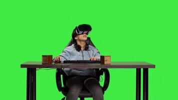 Pleased woman celebrating her games online success at computer, enjoys playing tournament with virtual reality interactive headset. Gamer feels happy against greenscreen backdrop. Camera B. photo