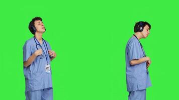Joyful nurse singing and dancing with wireless headphones on, listening to modern songs against greenscreen backdrop. Specialist enjoying favorite music and creating dance moves. Camera B. photo