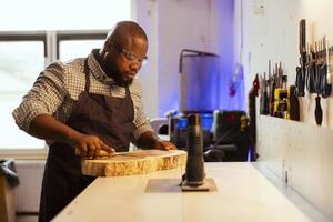 Carpenter wearing protection glasses doing last touches on piece of wood before using it for furniture assembly. Man doing woodworking process on lumber block, inspecting for damages photo