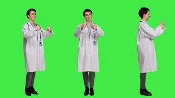 Cheerful medic doing thumbs up symbol against greenscreen backdrop, expresses positivity with like sign. General practitioner with coat giving approval and being satisfied with success. Camera B. photo