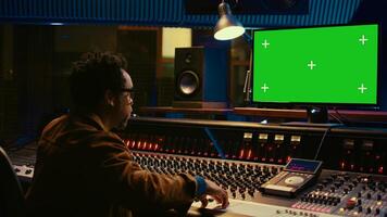 African american sound designer looks at greenscreen to record and edit tunes, uses mixing console to create new songs. Skilled technician works on processing and track recording. Camera B. photo