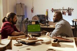 Smiling artisan and coworker comparing wood art piece with schematic displayed on isolated screen laptop. Woodworking experts in studio crosschecking wooden object with chroma key notebook blueprint photo