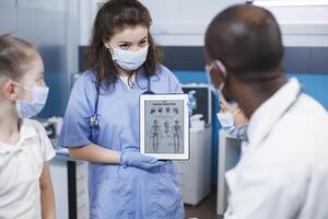 durante un visita, hueso médico y enfermero discutir el mejor médico alternativas para el paciente en el clínica oficina. un médico en antibacteriano protector engranaje muestra ortopédico información en un tableta. foto