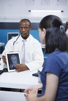 African American doctor uses a tablet to diagnose cervical vertebra damage and neck pain of female patient. Black man medical examiner in a lab coat advising woman of best possible treatments. photo