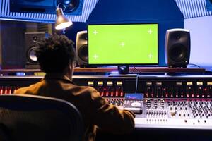 African american sound engineer works with greenscreen on computer in professional recording studio, mixing and mastering sounds on panel board. Young music producer pushing knobs and faders. photo