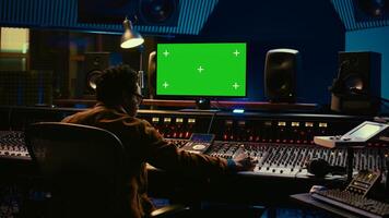 African american music producer working with greenscreen on pc, using mixing console to do mix and master in control room. Audio technician recording tracks with knobs and switchers. Camera B. photo