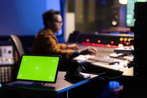 African american sound engineer editing music next to greenscreen display on gadget, operating on control room mixing console. Young producer pressing knobs and sliders in studio. photo