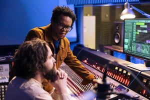 African american artist works with producer in professional studio, talking about adding new sound effects on tracks recorded. Young musician and technician editing songs with control panel desk. photo