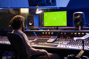 Sound designer working on mixing and mastering tracks with mockup display, using control panel board in professional studio. Skilled producer looks at greenscreen, editing music. photo