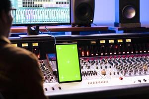 Singer and audio engineer collaborating on new hit song in studio, looks at mockup on gadget next to soundboard control desk panel board. Technician and artist recording new music, mixing console. photo